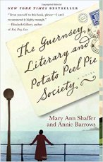 The Guernsey Literary and Potato Peel Pie Society y Mary Ann Shaffer (cover) Image: black text on a postcard....a woman dressed in a red coat stands at a railing overlooking the ocean